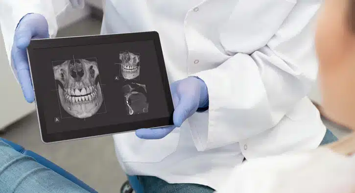 Dentist in gloves reviewing dental X-ray images on tablet in clinical setting.